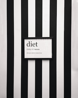 A framed definition of the word 'diet' displayed on a wall with vertical black and white stripes. The definition humorously states 'always starts tomorrow' beneath a phonetic spelling.