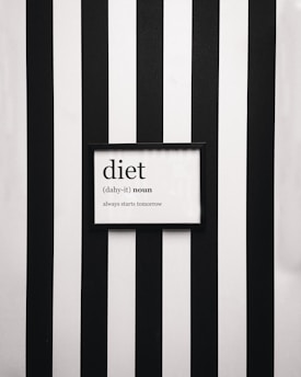 A framed definition of the word 'diet' displayed on a wall with vertical black and white stripes. The definition humorously states 'always starts tomorrow' beneath a phonetic spelling.