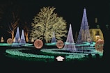 A night scene featuring a garden adorned with colorful holiday lights. Several cone-shaped trees and spherical ornaments are illuminated in vibrant colors, including blue, purple, and orange. The trees are wrapped in strings of lights, contributing to the festive and magical atmosphere. A pathway of green lights weaves through the display, leading the viewer's eye toward a large tree in the background, which is densely decorated with white lights. A building in the background is also partially visible, adding a sense of elegance to the scene.