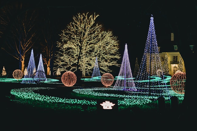 A night scene featuring a garden adorned with colorful holiday lights. Several cone-shaped trees and spherical ornaments are illuminated in vibrant colors, including blue, purple, and orange. The trees are wrapped in strings of lights, contributing to the festive and magical atmosphere. A pathway of green lights weaves through the display, leading the viewer's eye toward a large tree in the background, which is densely decorated with white lights. A building in the background is also partially visible, adding a sense of elegance to the scene.