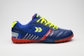 A colorful sports shoe with a blue body and red accents is displayed. It features a vibrant yellow lace and distinctive arrow design on the side. The shoe's sole is textured for grip, and the fabric includes a pattern with neon highlights.