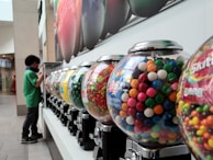 A row of colorful candy vending machines is displayed, each containing a variety of candies like gumballs and Skittles. A child stands to the side, seemingly interested in the machines. The setting appears to be indoors, possibly in a shopping mall, with bright overhead lighting and a large advertisement on the wall.