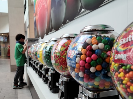 A row of colorful candy vending machines is displayed, each containing a variety of candies like gumballs and Skittles. A child stands to the side, seemingly interested in the machines. The setting appears to be indoors, possibly in a shopping mall, with bright overhead lighting and a large advertisement on the wall.