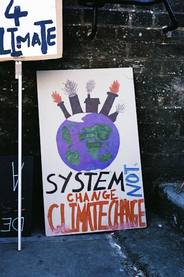 A protest sign with a drawing of Earth topped by factory smokestacks emitting smoke and flames, accompanied by the text 'SYSTEM CHANGE NOT CLIMATE CHANGE' in bold letters. The sign leans against a brick wall along with another partially visible sign.