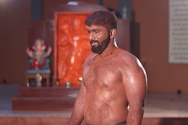 A muscular man with a beard stands confidently in an indoor space. His skin appears to be covered with a substance resembling mud or clay. In the background, there is a colorful statue, possibly of a deity, situated on a platform with a partially visible wall adorned with patterns or text.