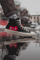 A close-up of a sneaker with a distinctive red heart and eyes design on the side, displayed in a reflective puddle. The sneaker is worn with plaid pants and stands out sharply against a blurred urban background.