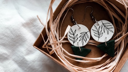 A pair of handcrafted earrings rests in a small cardboard box filled with straw-like material. The earrings feature circular white discs with black leaf patterns and dangling green triangular shapes.