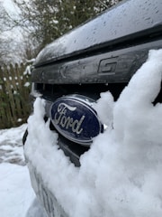 A close-up of a vehicle's front grill partially covered with snow, prominently displaying the Ford emblem. The background features a wooden fence and snow-covered ground.