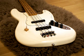 A white electric bass guitar lies on a shaggy, brown rug placed over a wooden floor. The guitar’s body is sleek with black control knobs and strings extending along the lighter wooden neck.