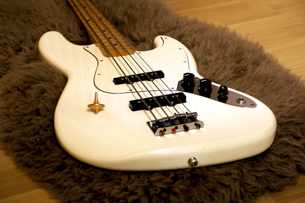 A white electric bass guitar lies on a shaggy, brown rug placed over a wooden floor. The guitar’s body is sleek with black control knobs and strings extending along the lighter wooden neck.