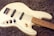 A cream-colored electric bass guitar rests on a textured, fluffy, brown surface. The guitar has a sleek finish with four strings, black pickups, and chrome volume knobs. Its fretboard is a light wood color, contrasting with the dark background.