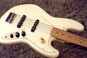 A cream-colored electric bass guitar rests on a textured, fluffy, brown surface. The guitar has a sleek finish with four strings, black pickups, and chrome volume knobs. Its fretboard is a light wood color, contrasting with the dark background.