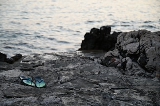 A pair of blue flip-flops rests on weathered, rocky terrain near a body of water. The surface is rough and uneven with a mix of dark and light gray stones. Waves gently ripple in the background, indicating a calm, coastal setting.