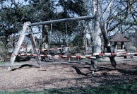 An outdoor playground swing structure with red and white caution tape wrapped around it, indicating it is closed or off-limits. The swing set is made of wood and metal, standing on a ground covered with wood chips. Surrounding the area are tall trees and a small wooden shelter is visible in the background.