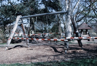 An outdoor playground swing structure with red and white caution tape wrapped around it, indicating it is closed or off-limits. The swing set is made of wood and metal, standing on a ground covered with wood chips. Surrounding the area are tall trees and a small wooden shelter is visible in the background.