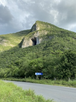 A large forest-covered hill or mountain with a noticeable cave opening near the top. The sky is mostly cloudy with patches of sunlight illuminating parts of the hill. Below, there is a road with a blue road sign that reads 'CUEVA DE LOS MURCIÉLAGOS'. Surrounding the road and hill are dense, green trees and bushes.