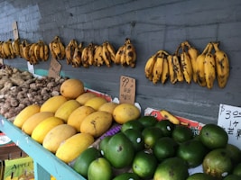 A variety of fruits and vegetables are displayed on a market stall. Bananas hang from the wall above, while below, there are piles of yams, yellow melons, and green mangos. Small signs with prices and some text in an Asian script are placed among the produce.