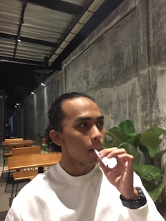 A person sitting indoors, holding a vape pen near their mouth. The setting includes a row of wooden tables and chairs, a textured concrete wall, and some green plants. The person is wearing a white shirt and a wristwatch.