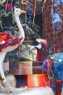 A festive holiday display featuring two large, decorative birds with Christmas ornaments and fir branches. The birds are adorned with red and gold sequins and beads. Behind them, strings of red and blue baubles hang from a Christmas tree, situated against a brick wall backdrop. Wrapped gift boxes and shiny decorations complete the scene, adding a festive and luxurious touch.