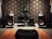 A cozy room featuring an elaborate audio setup with large speakers, audio equipment, and a decorative pattern on the wall. A leafy potted plant adds a touch of greenery. An orange shag rug and a black cushioned seat are positioned in the foreground.