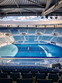 A large, empty tennis stadium with numerous blue seats surrounding a central blue court. Two people are visible on the court, one standing on each side of the net. The stadium has a modern design with a partially covered ceiling.