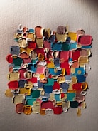 Abstract painting composed of thick, textured squares and rectangles in various bright colors. The brushstrokes are layered and visible, creating a 3D effect against a canvas background.