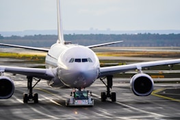 An airplane is being towed on the tarmac of an airport by a small vehicle. The aircraft's large engines and wings are prominently visible. The landscape in the background is composed of trees and a cloudy sky.
