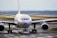 An airplane is being towed on the tarmac of an airport by a small vehicle. The aircraft's large engines and wings are prominently visible. The landscape in the background is composed of trees and a cloudy sky.