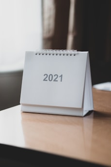 A desk calendar for the year 2021 is placed on a wooden table. The calendar has a simple, minimalistic design with a spiral binding at the top, and the number 2021 is printed in large, gray text on a white background.