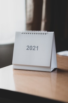 A desk calendar for the year 2021 is placed on a wooden table. The calendar has a simple, minimalistic design with a spiral binding at the top, and the number 2021 is printed in large, gray text on a white background.