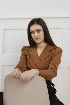 A person with long dark hair is sitting on a light-colored chair, wearing a brown blazer with shoulder pads. The background features a white paneled wall, creating a minimalistic and clean environment.