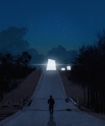 A mysterious, illuminated geometric structure is situated at the end of a long, deserted road lined with trees under a starry night sky. The scene has a surreal, almost otherworldly atmosphere, with a solitary figure standing near a stop sign in the foreground.