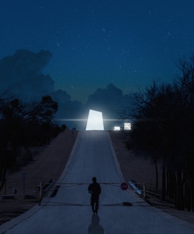 A mysterious, illuminated geometric structure is situated at the end of a long, deserted road lined with trees under a starry night sky. The scene has a surreal, almost otherworldly atmosphere, with a solitary figure standing near a stop sign in the foreground.
