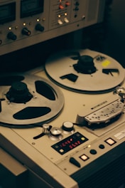 A vintage reel-to-reel tape recorder is shown with two large reels. The device has multiple knobs, switches, and meters, indicating its functionality for audio recording or playback. The design is industrial and nostalgic, evoking the analog era of audio technology.