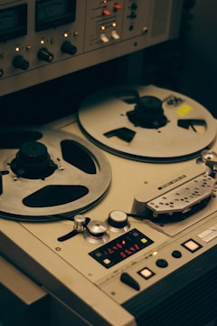 A vintage reel-to-reel tape recorder is shown with two large reels. The device has multiple knobs, switches, and meters, indicating its functionality for audio recording or playback. The design is industrial and nostalgic, evoking the analog era of audio technology.