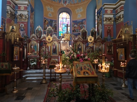 An ornate interior of a church adorned with religious icons and paintings. The walls are richly decorated with colorful frescoes and iconography. Candles are lit throughout the space, providing a warm and solemn glow. A stained glass window at the end of the room adds vibrant colors to the sacred setting. Ornate wooden structures hold numerous icons, while flowers are displayed prominently at the altar.