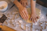 Hands of an adult and child working together to roll out dough on a floured surface. Various cookie cutters in different shapes like stars and trees are scattered around. There is also a white bowl filled with flour and a rolling pin nearby.