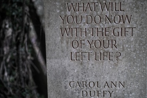 A gray stone slab engraved with the words, 'WHAT WILL YOU DO NOW WITH THE GIFT OF YOUR LEFT LIFE?' followed by the name 'CAROL ANN DUFFY'. The surface has a textured pattern, and the background features blurred greenery.