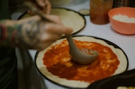 A chef spreading creamy sauce over a pizza dough in a kitchen.