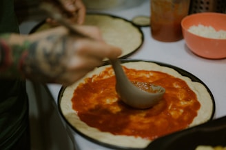 A chef spreading creamy sauce over a pizza dough in a kitchen.