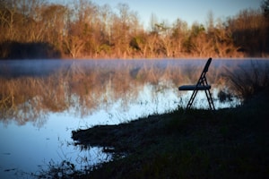 A serene outdoor scene featuring a tranquil lake reflecting the bare trees of autumn. A single folding chair is positioned at the edge of the water, creating a sense of solitude. Mist hovers over the water, adding a mystical atmosphere.