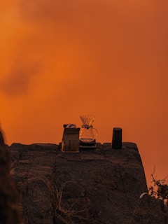 On a rocky surface set against a vibrant orange sky, a pour-over coffee setup is arranged, featuring a glass carafe with a filter, a bag of coffee, and a tumbler.