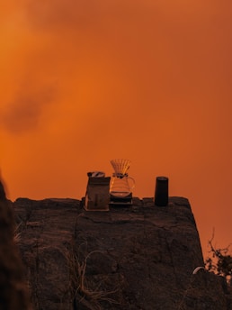 On a rocky surface set against a vibrant orange sky, a pour-over coffee setup is arranged, featuring a glass carafe with a filter, a bag of coffee, and a tumbler.
