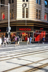 A busy urban street corner with a prominent store named Mecca. The building features large windows and a beige facade. People are walking on the sidewalk, some carrying shopping bags, indicating a shopping district. The street has tram tracks and a one-way sign, suggesting city transportation infrastructure.