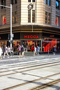 A busy urban street corner with a prominent store named Mecca. The building features large windows and a beige facade. People are walking on the sidewalk, some carrying shopping bags, indicating a shopping district. The street has tram tracks and a one-way sign, suggesting city transportation infrastructure.