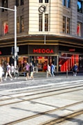 A busy urban street corner with a prominent store named Mecca. The building features large windows and a beige facade. People are walking on the sidewalk, some carrying shopping bags, indicating a shopping district. The street has tram tracks and a one-way sign, suggesting city transportation infrastructure.