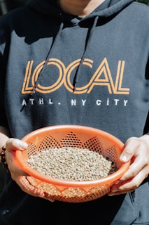A person is holding an orange basket filled with green coffee beans. The individual is wearing a navy blue hoodie with the word 'LOCAL' printed in bold letters across the chest, accompanied by smaller text reading 'ATHL. NY CITY'.