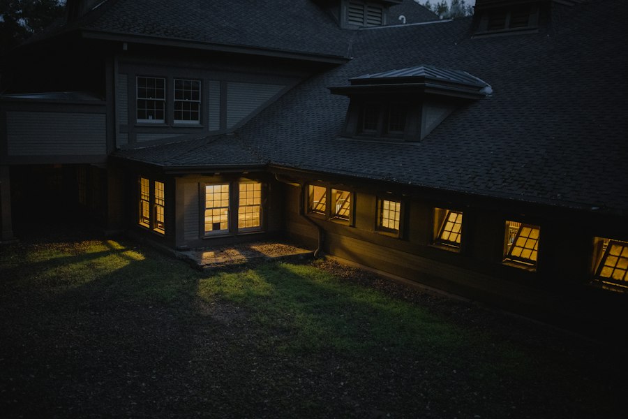 Shingled house with dormers and warm lit windows above a damp lawn at twilight