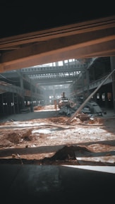 A large industrial warehouse under construction or renovation. Sunlight streams through gaps in the structure, casting shadows on the dirt and scattered construction materials. Steel beams and partially constructed walls create a complex geometric pattern. There is an excavator in the center, indicating ongoing work.