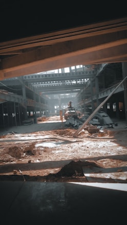 A large industrial warehouse under construction or renovation. Sunlight streams through gaps in the structure, casting shadows on the dirt and scattered construction materials. Steel beams and partially constructed walls create a complex geometric pattern. There is an excavator in the center, indicating ongoing work.